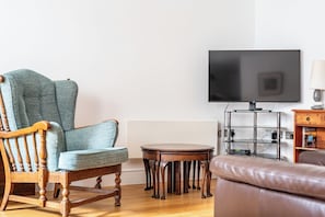 Ein Wohnzimmer mit einem Holzstuhl, einem kleinen runden Tisch, einem an der Wand befestigten Fernseher und einer Lampe auf einem Beistelltisch.