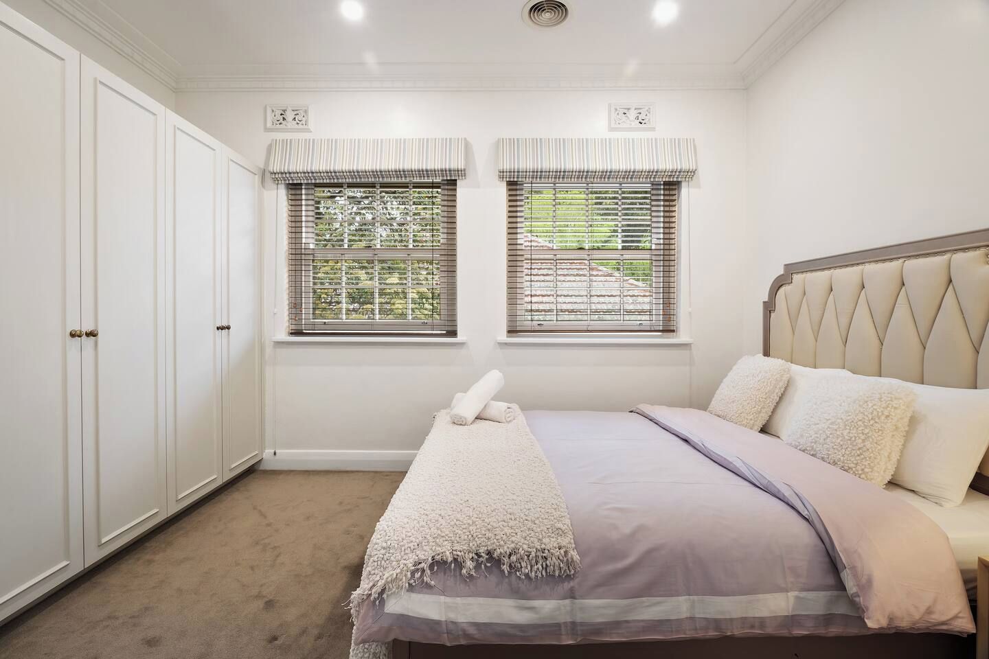 A bedroom with a bed, two windows with blinds, a wardrobe, and a ceiling light.