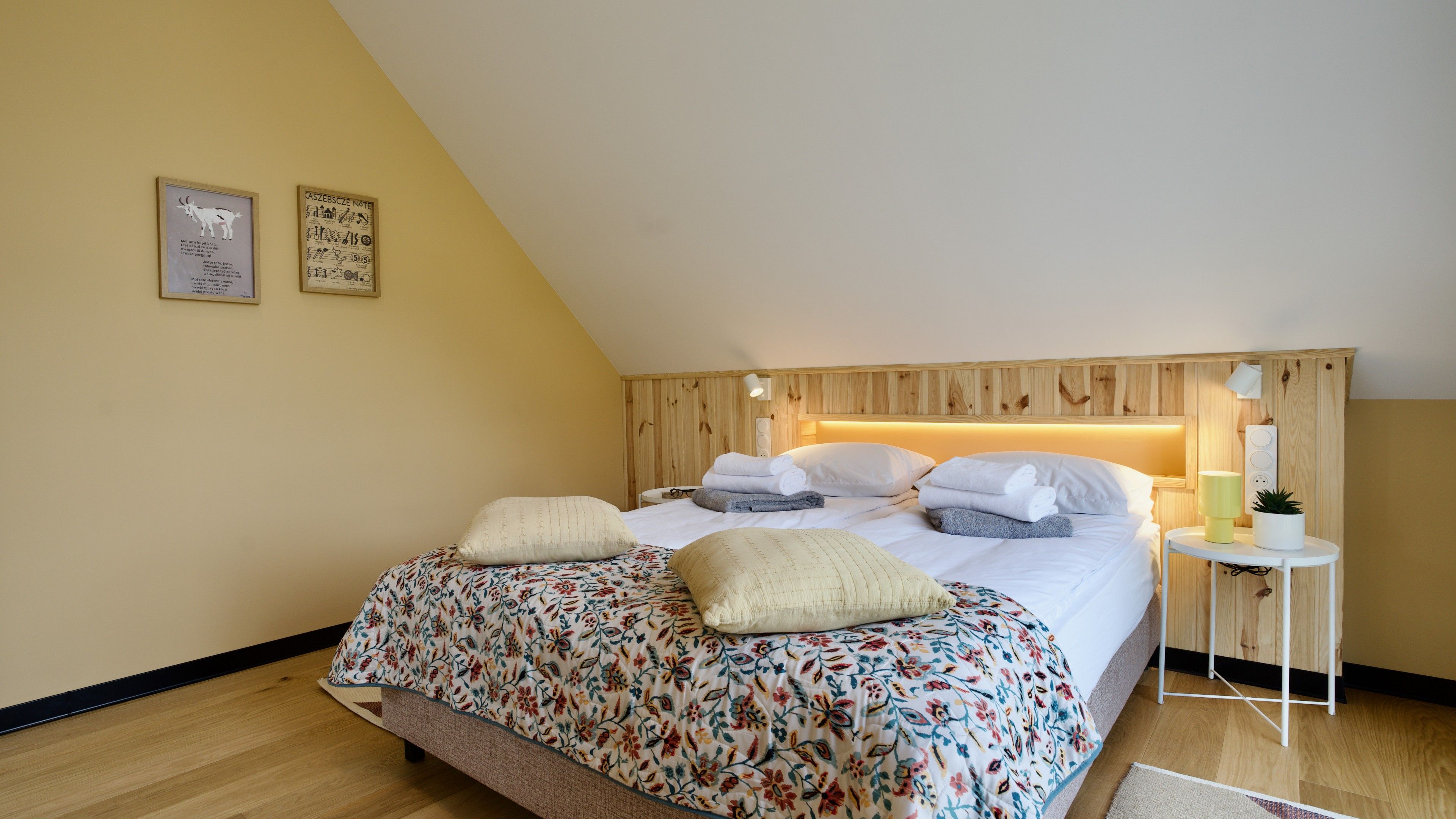A bedroom with a sloped ceiling, a bed with floral bedding, a bedside table with a lamp, and a small plant.