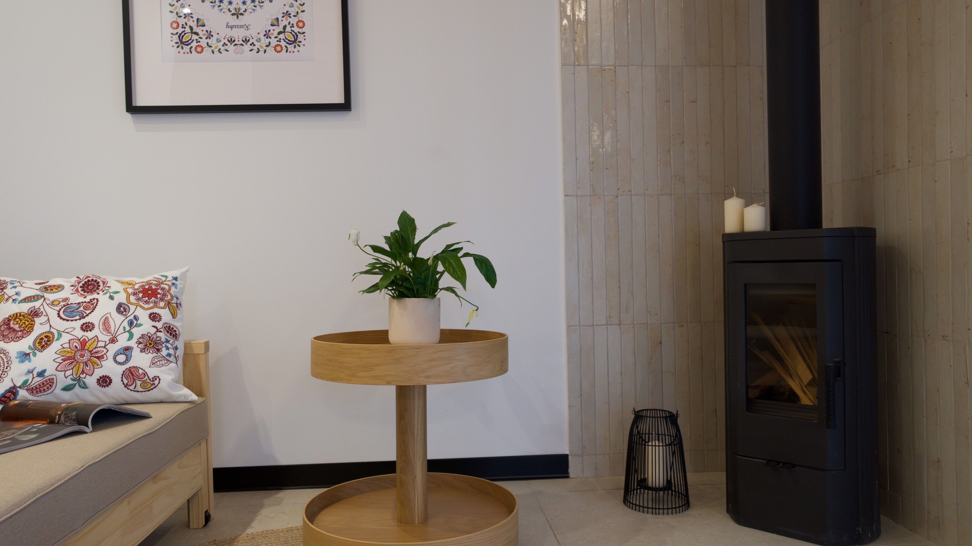 A room with a round wooden table with a potted plant, a sofa with a patterned cushion, and a black wood-burning stove.