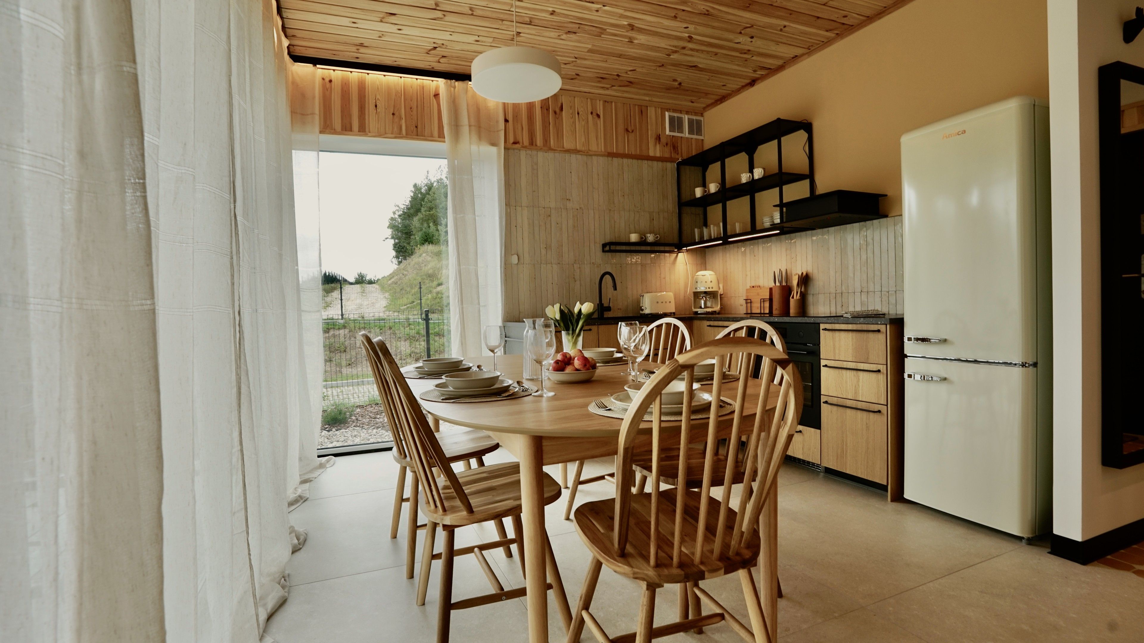Et moderne køkken med træskabe, et hvidt køleskab samt spiseplads med træstole og et bord dækket til fire personer.