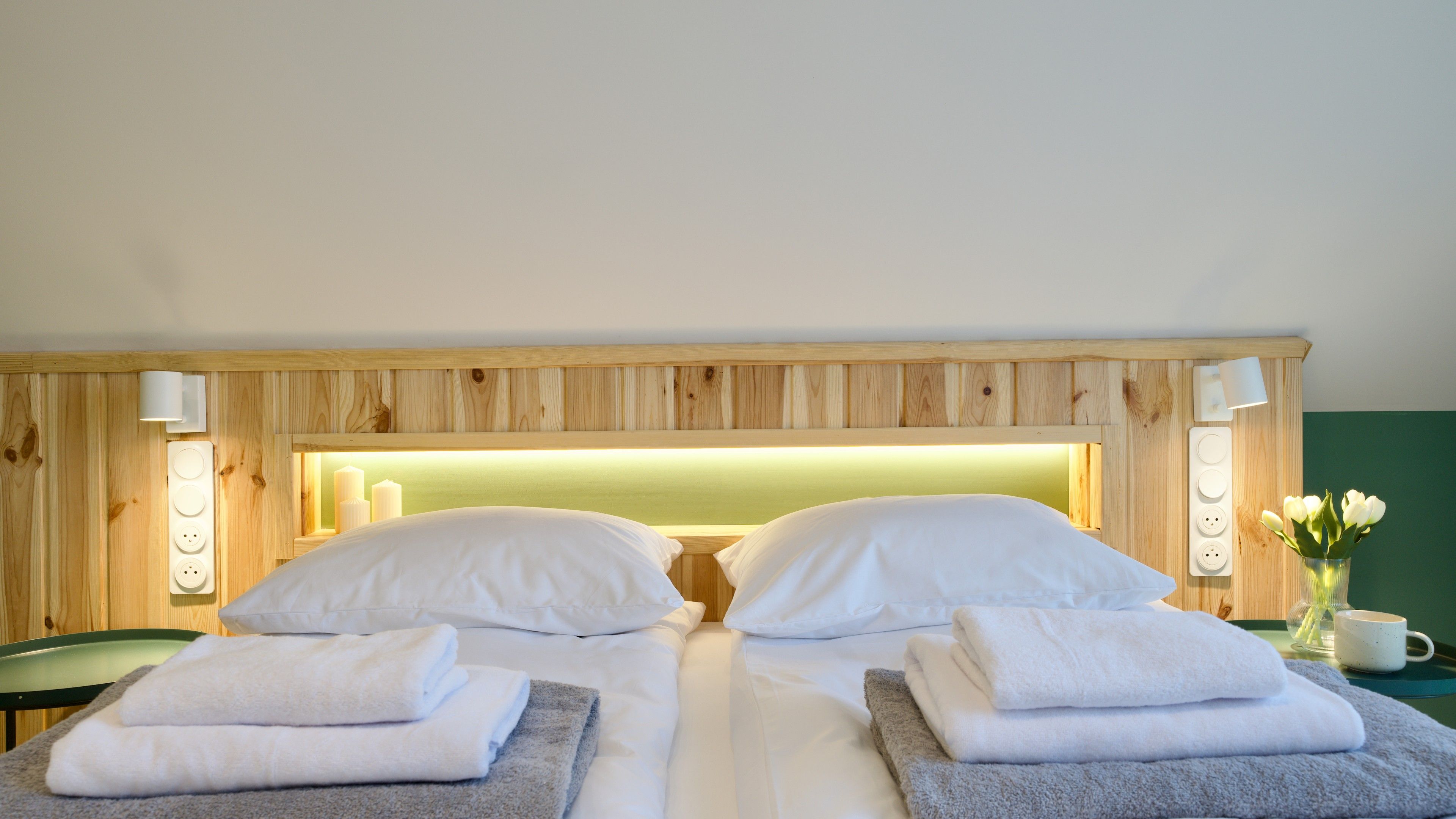 Ein Hotelzimmer mit einem hölzernen Kopfteil, zwei weißen Kissen, einem ordentlich bezogenen Bett mit weißer Bettwäsche, einem grünen Sessel und einem kleinen Tisch mit Blumen in einer Vase und einer Tasse.