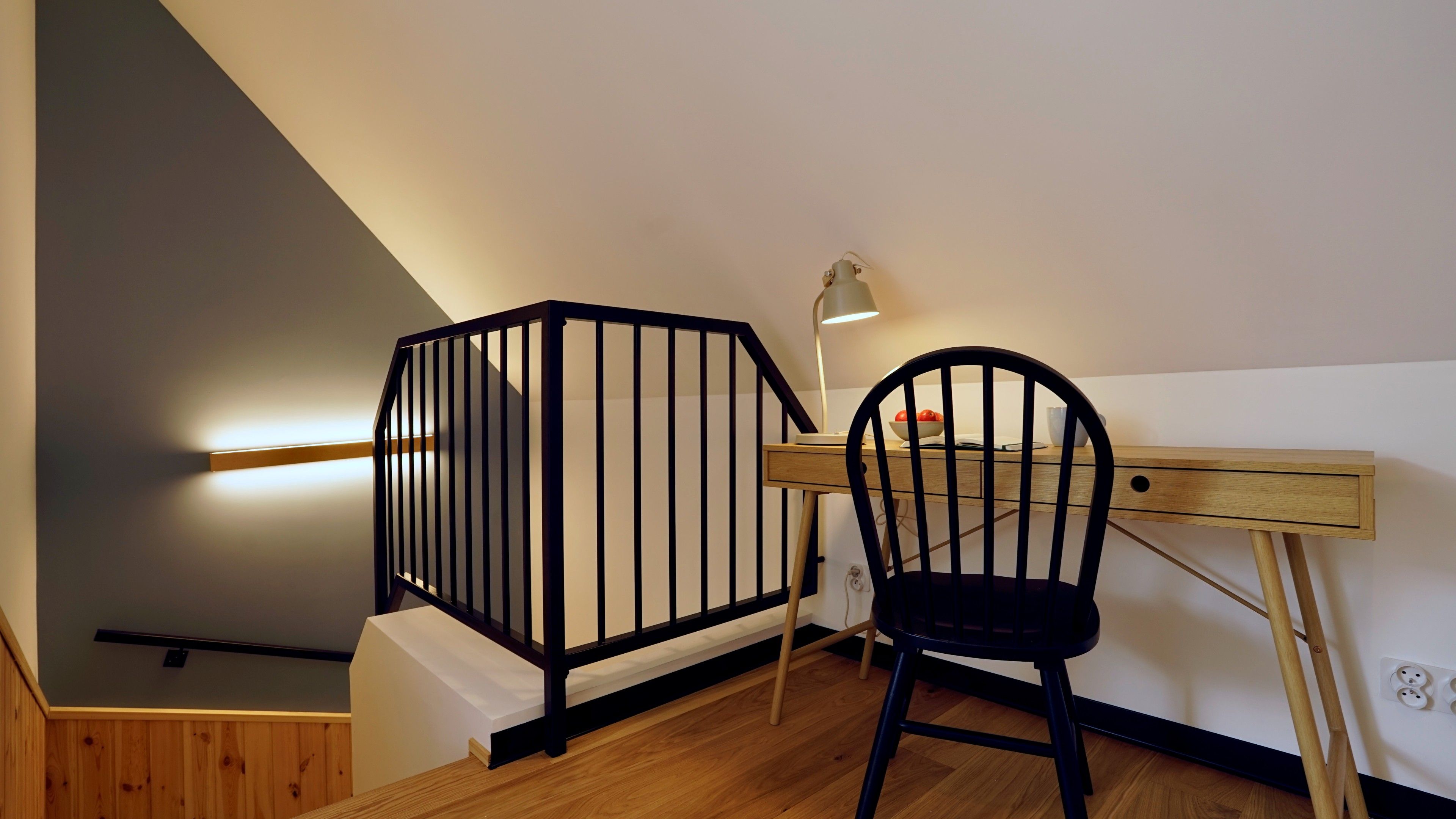 Une chambre moderne avec un bureau en bois, une chaise noire, une rambarde en métal noir et une lampe fixée au mur.