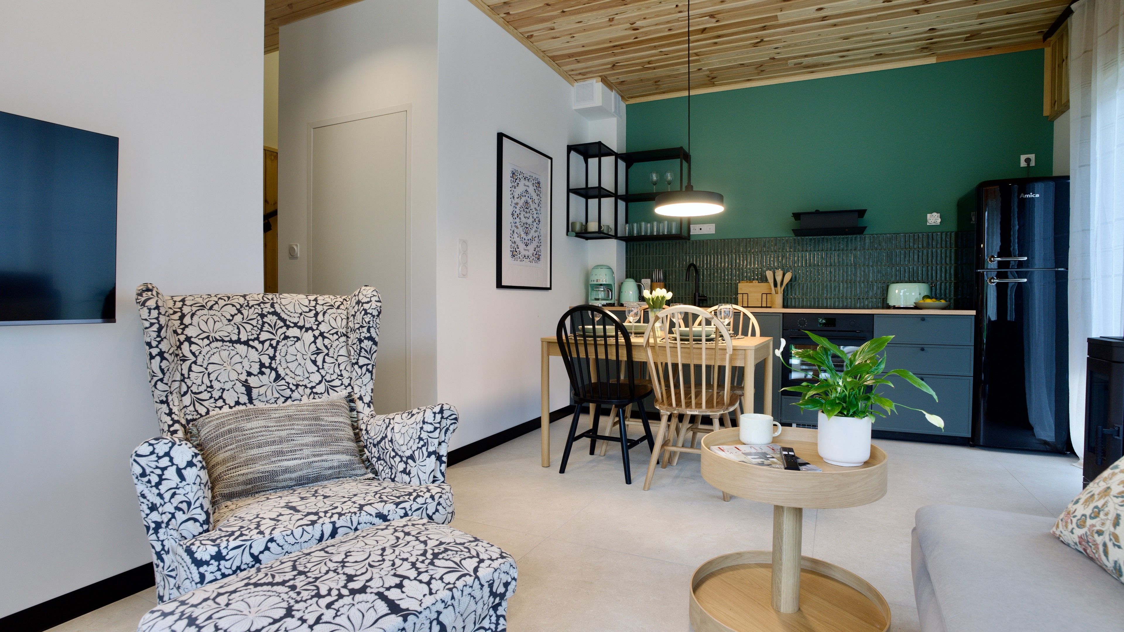 Una sala de estar con un sillón estampado, una mesa de comedor con sillas, una mesita de centro con una planta, y una cocina con un refrigerador y armarios.