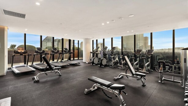 A modern gym with treadmills, elliptical machines, and weight stations, featuring large windows and a city view. - Sky One 3BR, 4 Min Walk to Box Hill Station - Apartment (Box Hill)