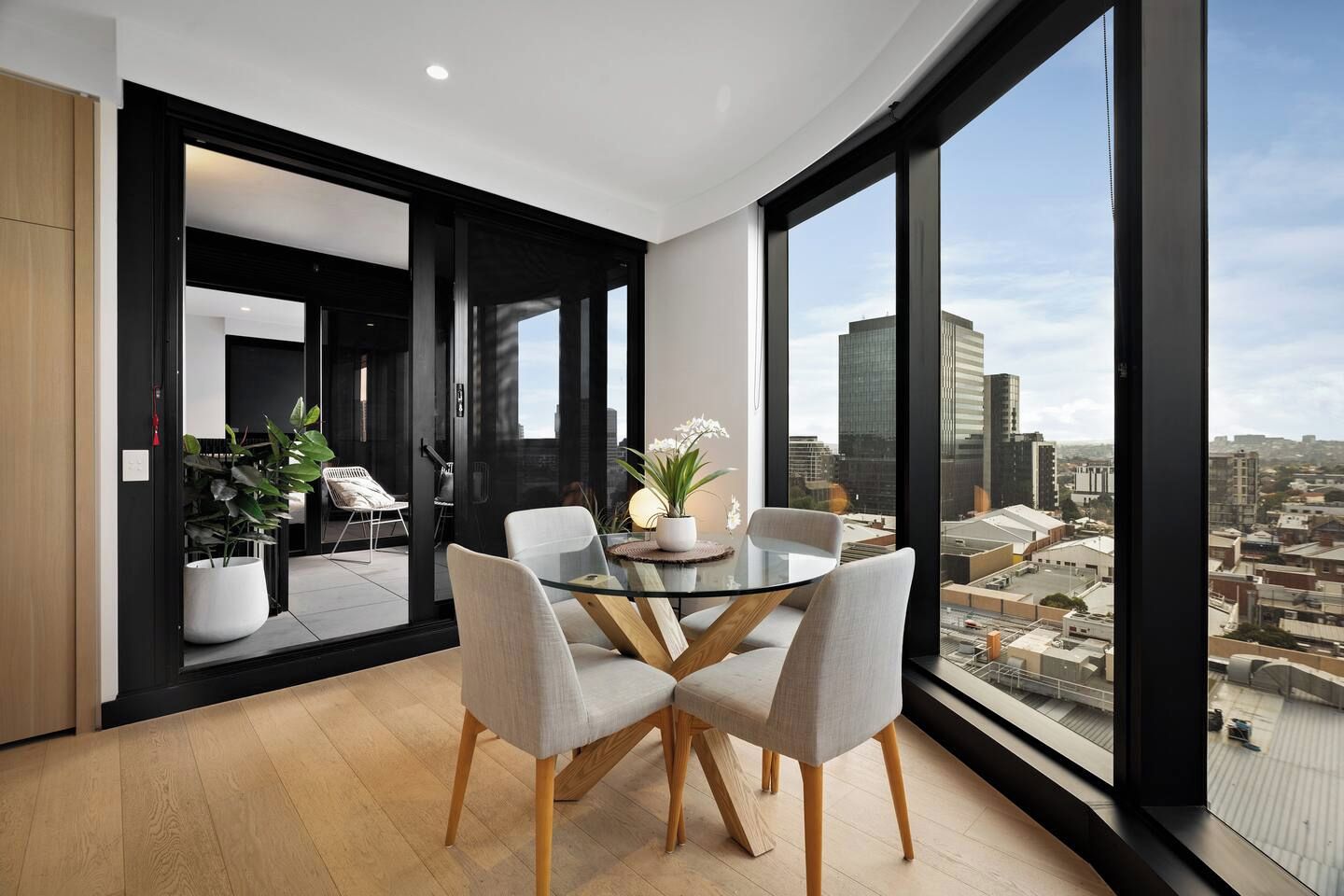 A modern dining area with a glass table and chairs, a potted plant, and a city view through large windows.