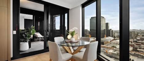 A modern dining area with a glass table and chairs, a potted plant, and a city view through large windows.