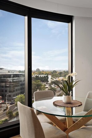 A modern dining area with a glass table and chairs, a potted plant, and a view of a cityscape through large windows. - Sky One 3BR, 4 Min Walk to Box Hill Station - Apartment (Box Hill)