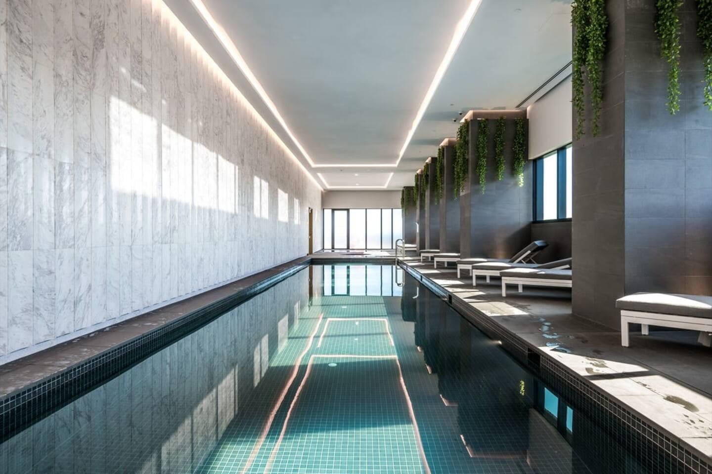 A modern indoor swimming pool with a long, narrow design, surrounded by seating areas and a tiled floor.