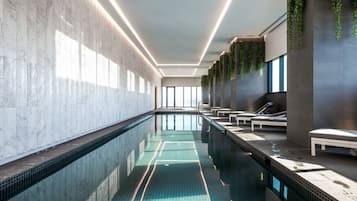 A modern indoor swimming pool with a long, narrow design, surrounded by seating areas and a tiled floor.