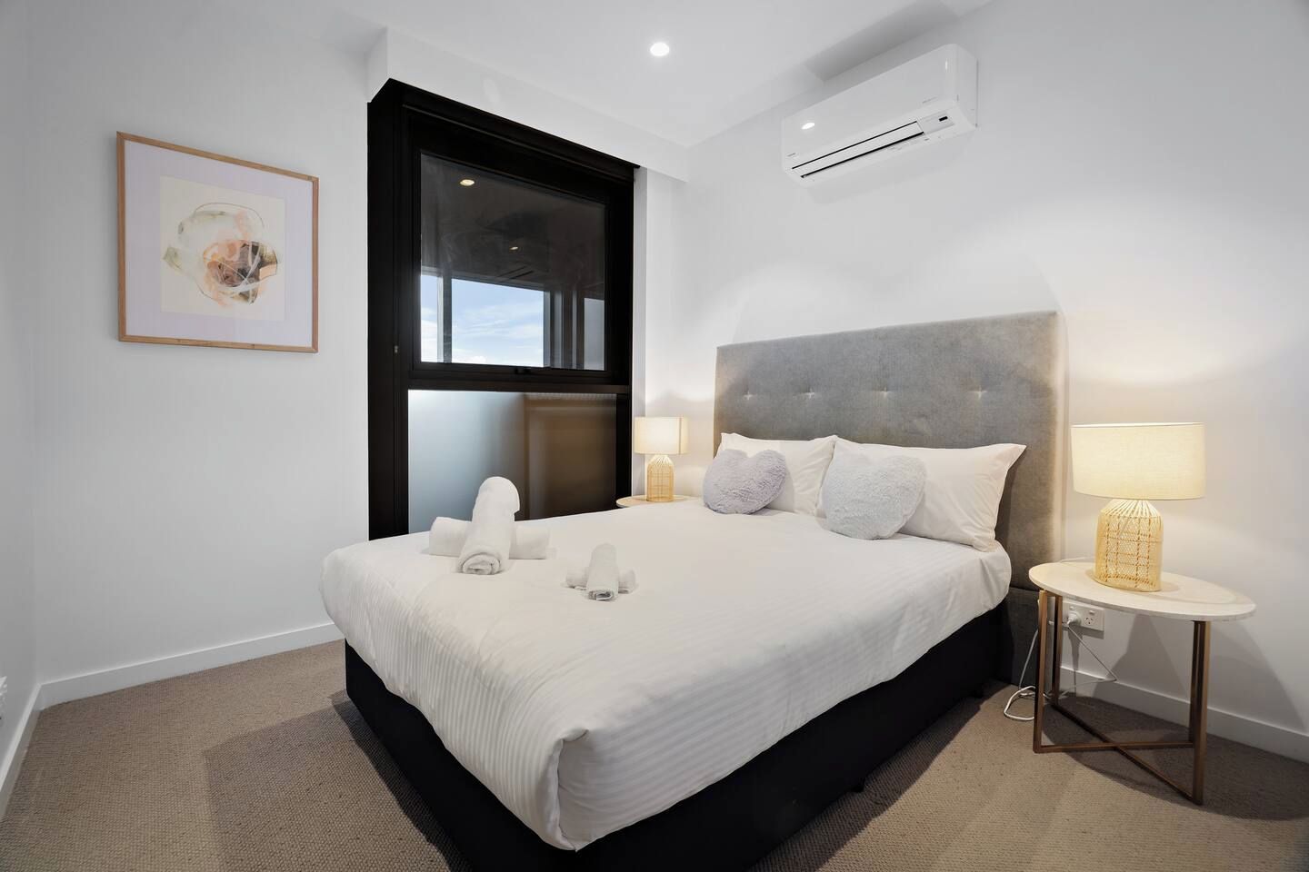A hotel room with a large bed, two bedside lamps, a window, and an air conditioning unit.