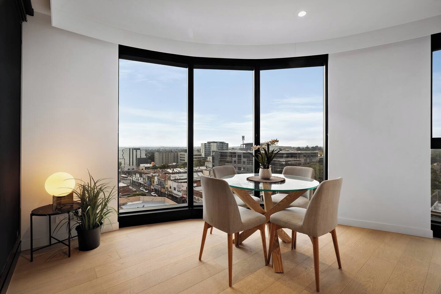Ein moderner Essbereich mit einem Glastisch und Stühlen, einer Küstpflanze und Blick auf eine Stadtlandschaft durch große Fenster.