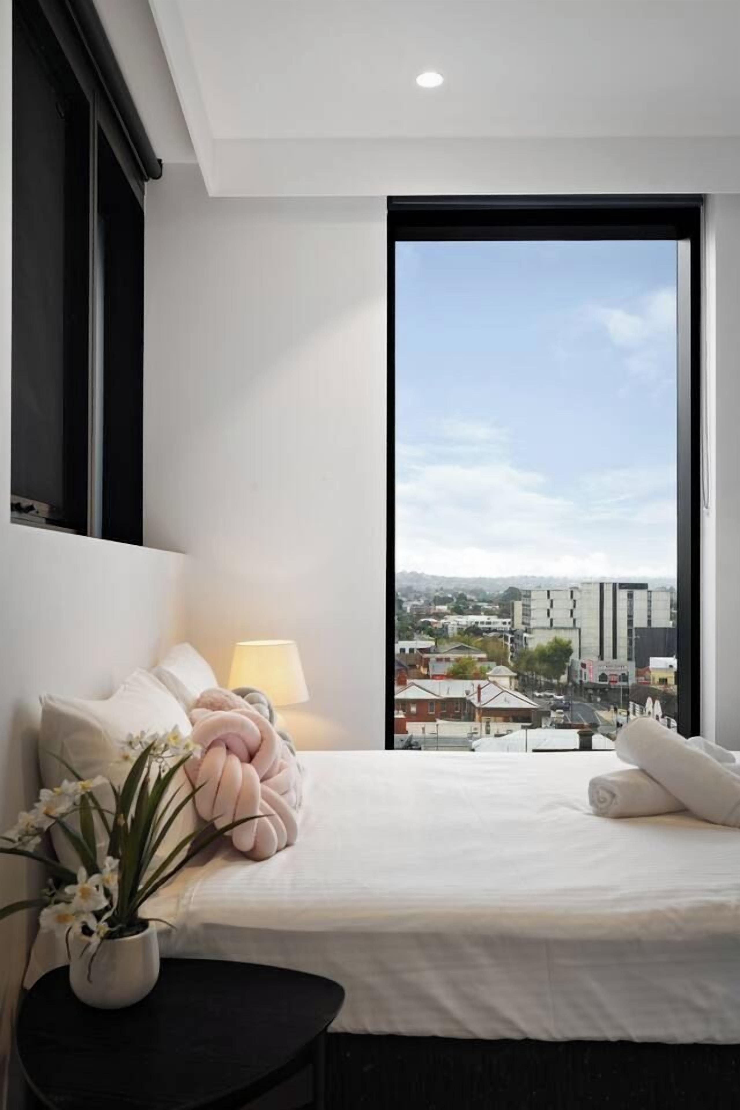 A bedroom with a large window, a bed with pillows, a bedside lamp, and a potted plant.