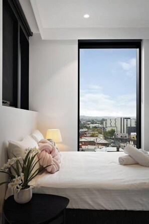 A bedroom with a large window, a bed with pillows, a bedside lamp, and a potted plant. - Sky One 3BR, 4 Min Walk to Box Hill Station - Apartment (Box Hill)