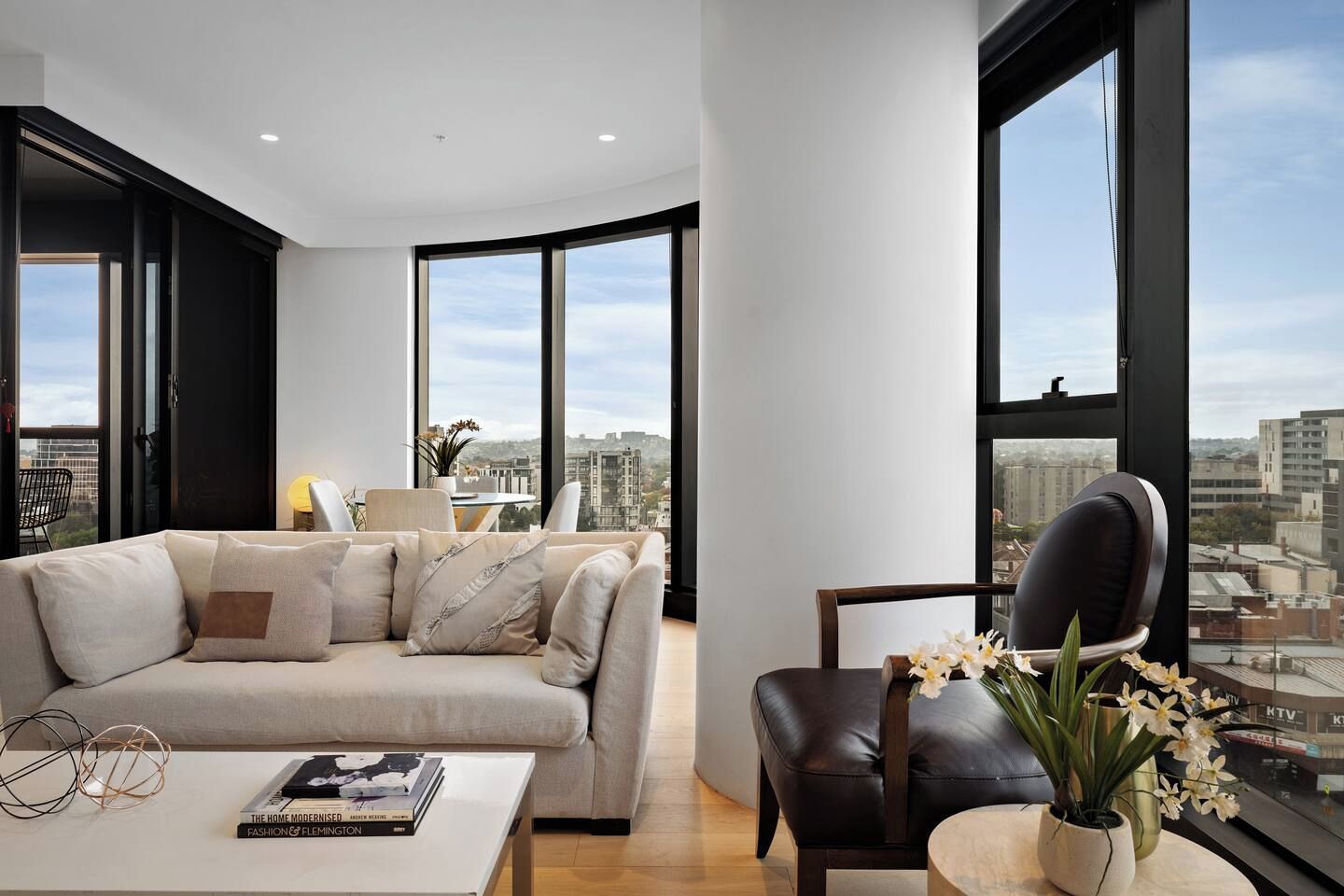A modern living room with a sofa, a coffee table with a book, a chair, and a view of the city through large windows.