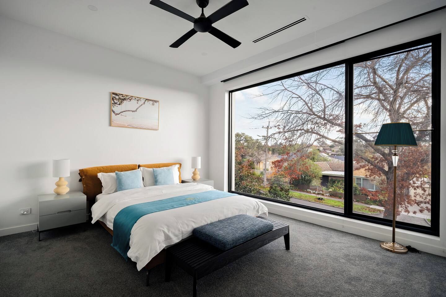 A bedroom with a large window, a bed with a blue and white bedspread, a bedside table with a lamp, and a ceiling fan.