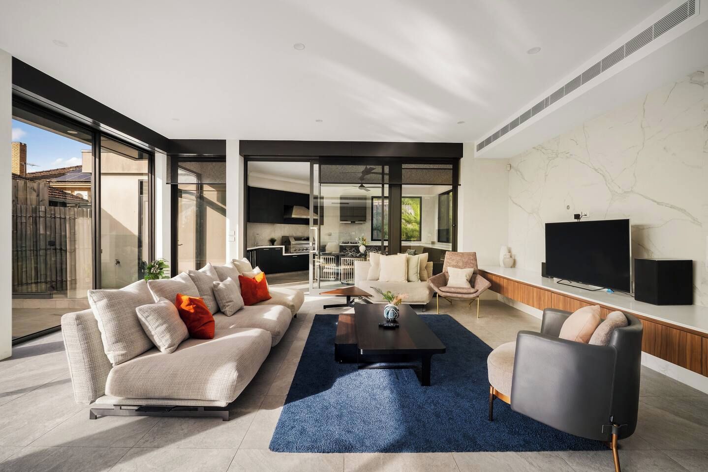 A modern living room with a sofa, coffee table, and armchairs, a television, and a kitchen in the background.