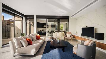 A modern living room with a sofa, coffee table, and armchairs, a television, and a kitchen in the background.