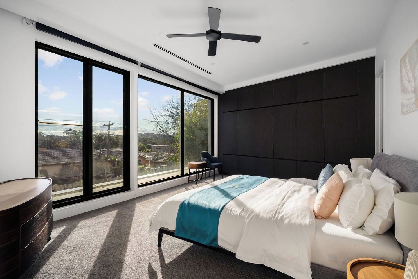A modern bedroom with a large bed, a ceiling fan, and a view of the outdoors through floor-to-ceiling windows.