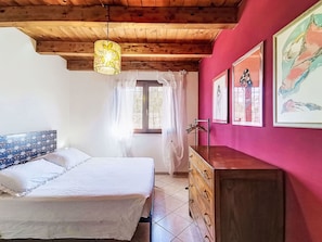 Ein Schlafzimmer mit einem Bett, einer Holzkommode, einem Fenster mit Vorhängen und Kunstwerken an der Wand.
