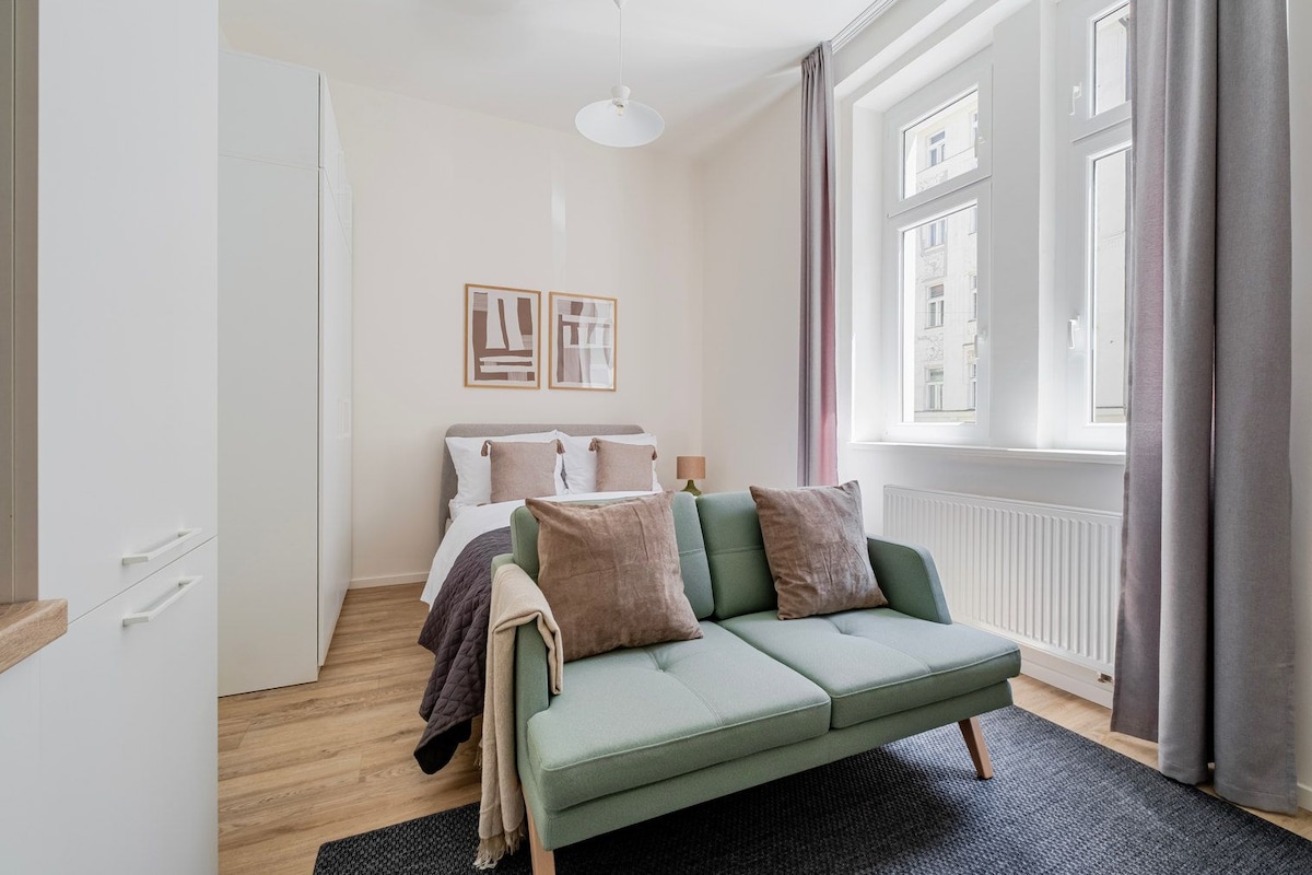 A modern bedroom with a green sofa, a bed with white and brown bedding, a white wardrobe, and a window with curtains.