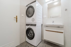 A compact laundry room with a front-loading washer and dryer, a sink with a mirror above it, and a door with a handle. - Letná Residence by Blueground (Prague)