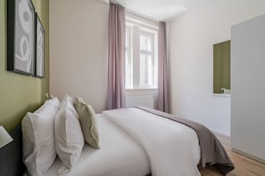 A bedroom with a bed, pillows, a window with curtains, a radiator, and a mirror on the wall. - Letná Residence by Blueground (Prague)