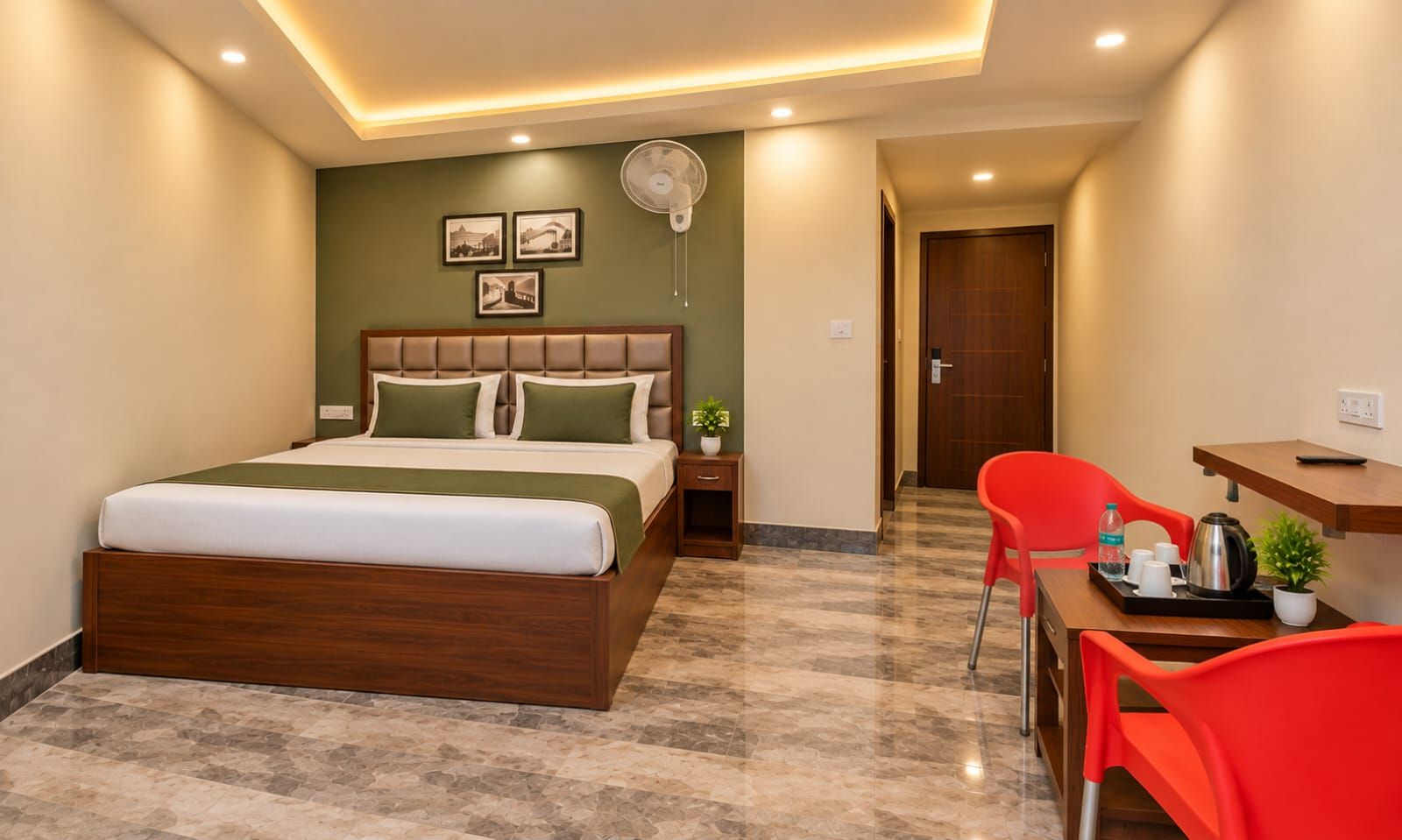 Una habitación de hotel con cama, escritorio, silla, ventilador y una pequeña planta.