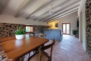 A dining area with a wooden table and chairs, a potted plant, a stone wall, and a sofa in the background.