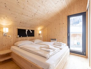 A wooden cabin bedroom with a bed, two wall-mounted lights, a black and white painting, and a window with a view of the outside. - One-bedroom Cabin (Sankt Georgen am Kreischberg)