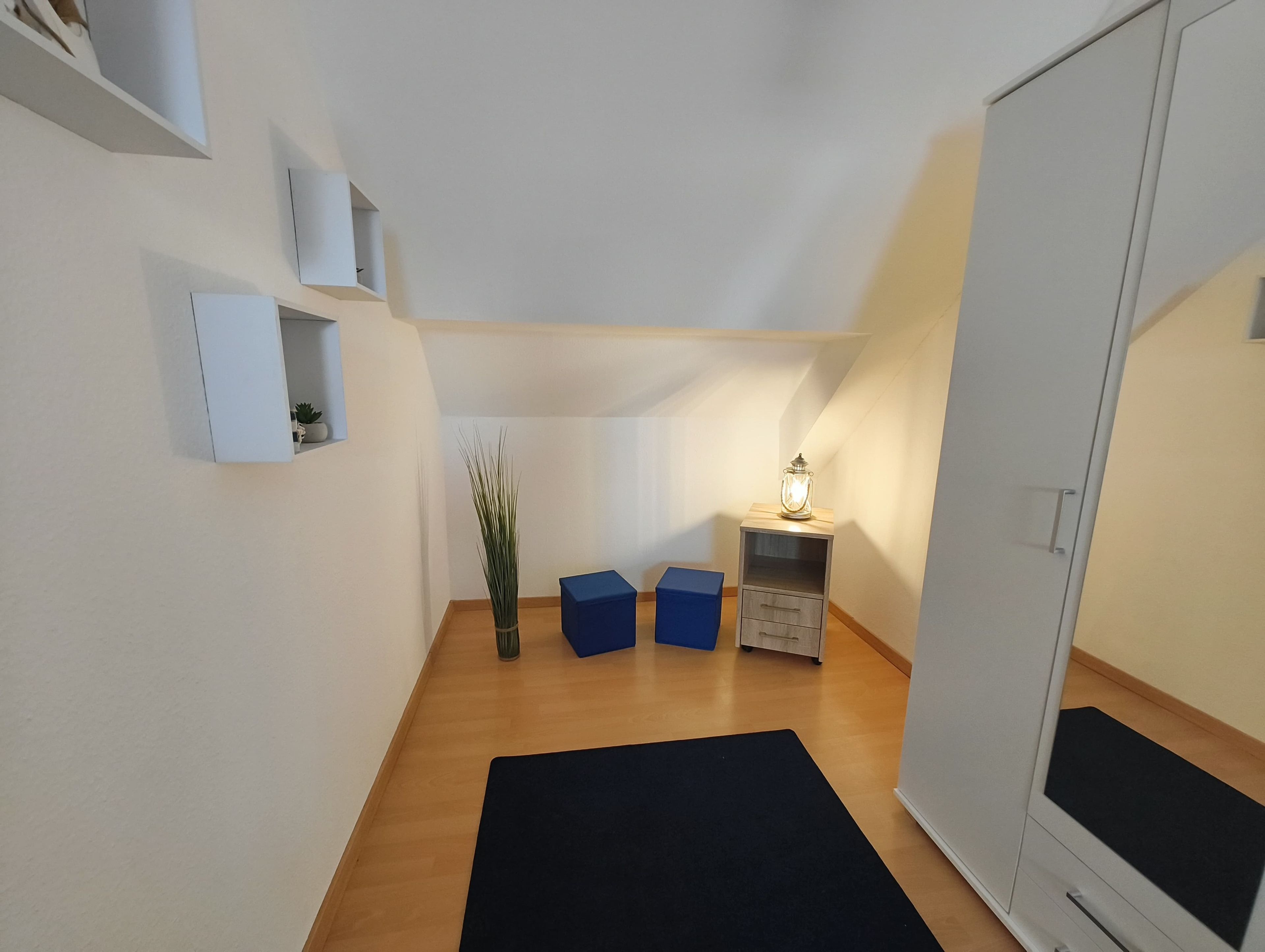 Um corredor estreito com piso de madeira, tapete preto, caixa de armazenamento azul, mesa pequena com luminária e guarda-roupa branco.