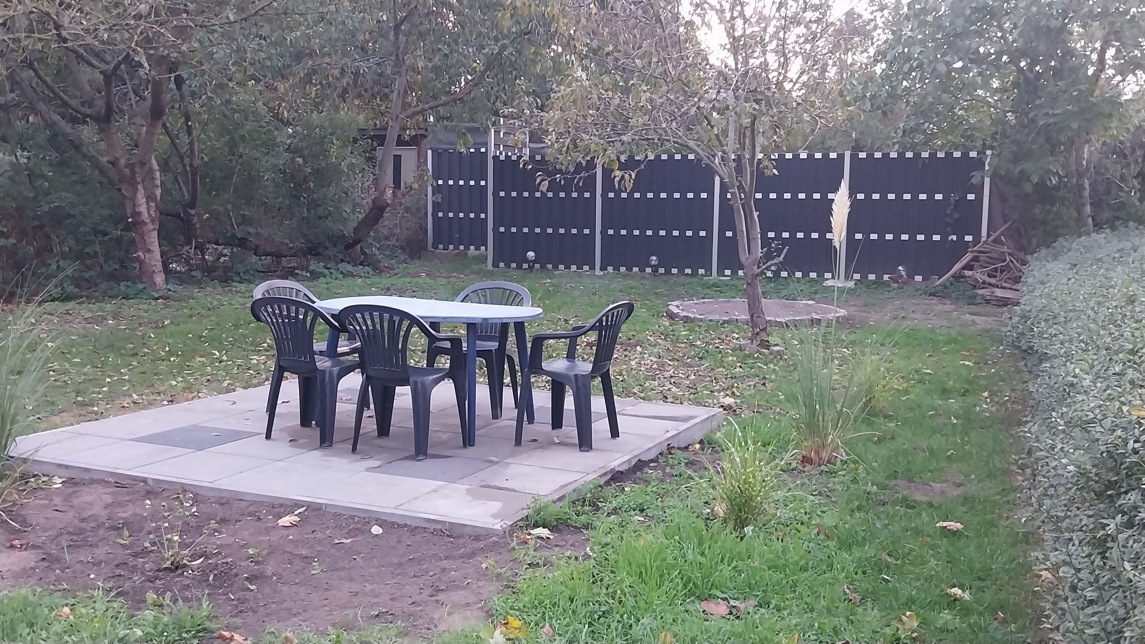 Une terrasse extérieure avec une table et des chaises, entourée de verdure et d’une clôture.