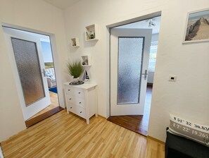 A hallway with wooden flooring, white walls, and a door with frosted glass. There are small shelves with decorative items and a plant on the wall.
