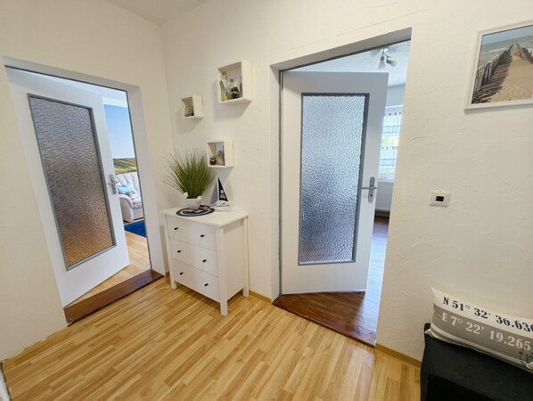 Un pasillo con piso de madera, paredes blancas y una puerta con vidrio esmerilado. Hay pequeñas estanterías con objetos decorativos y una planta en la pared.