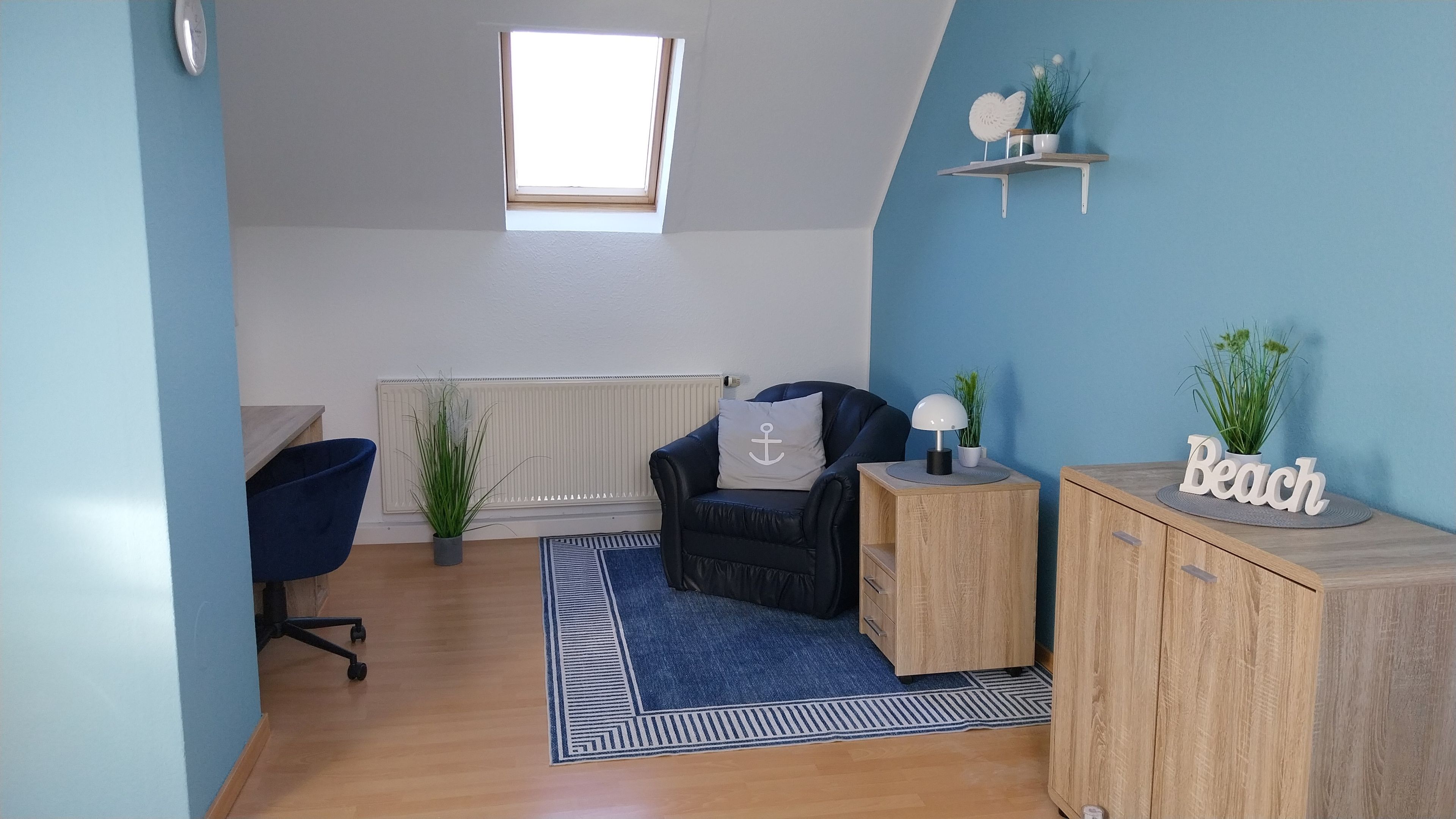 A room with a blue wall, a black leather chair, a wooden cabinet, a small table with a lamp, and a shelf with plants and a sign.