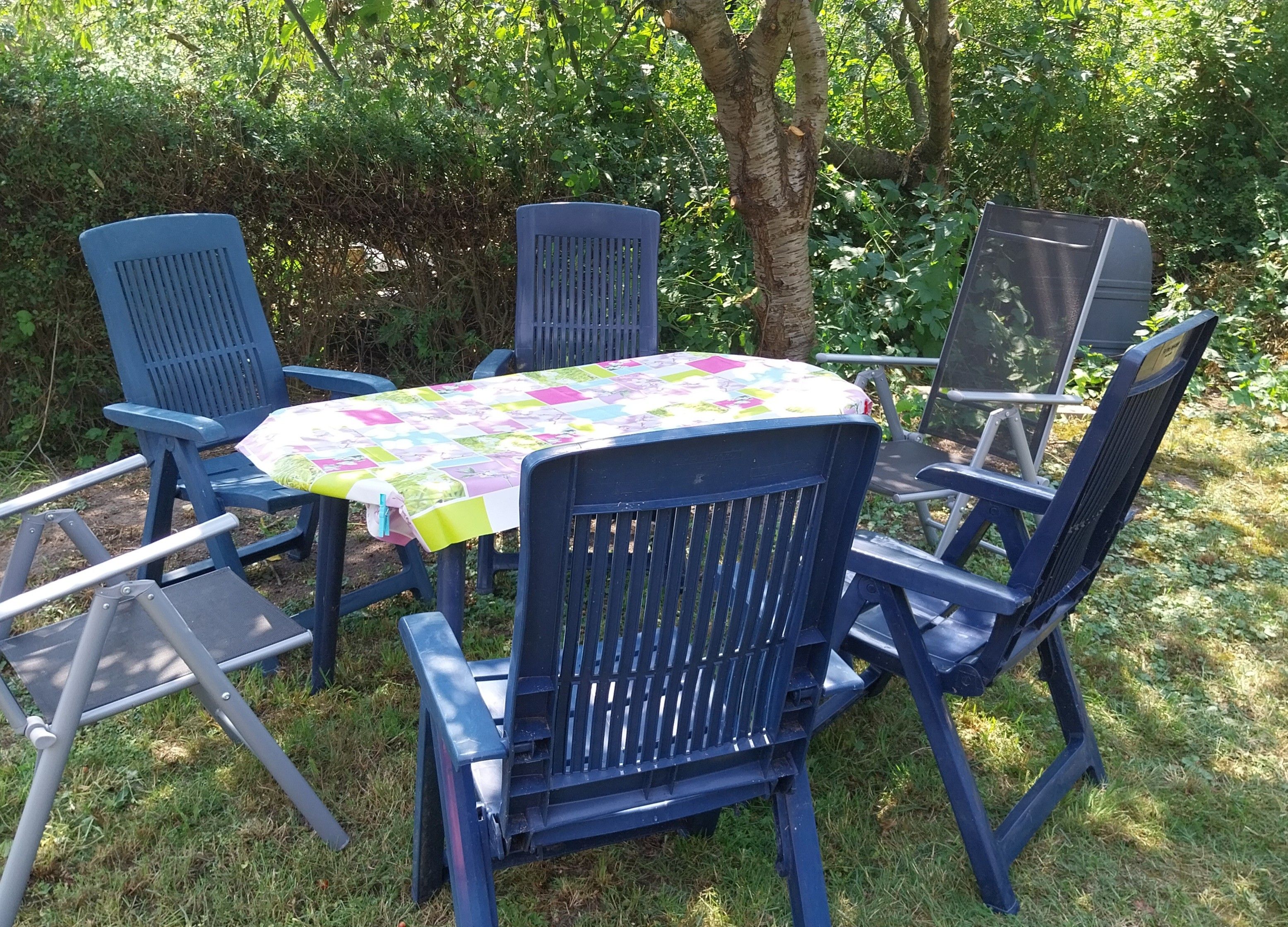 A set of blue chairs and a table with a colorful tablecloth, arranged outdoors with greenery in the background.