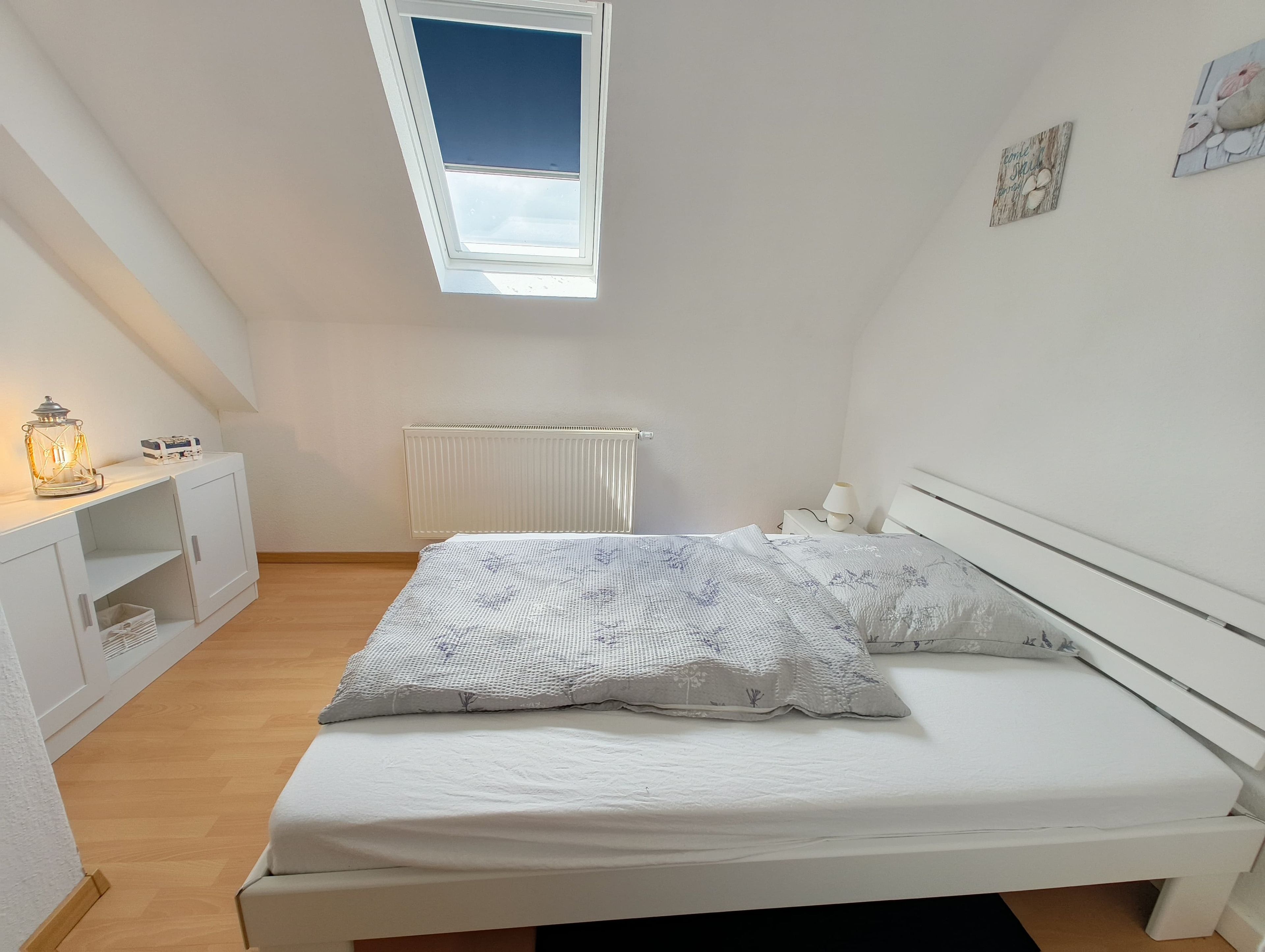 Une chambre à coucher comprenant un lit, une table de chevet, un radiateur, une fenêtre et un tableau accroché au mur.