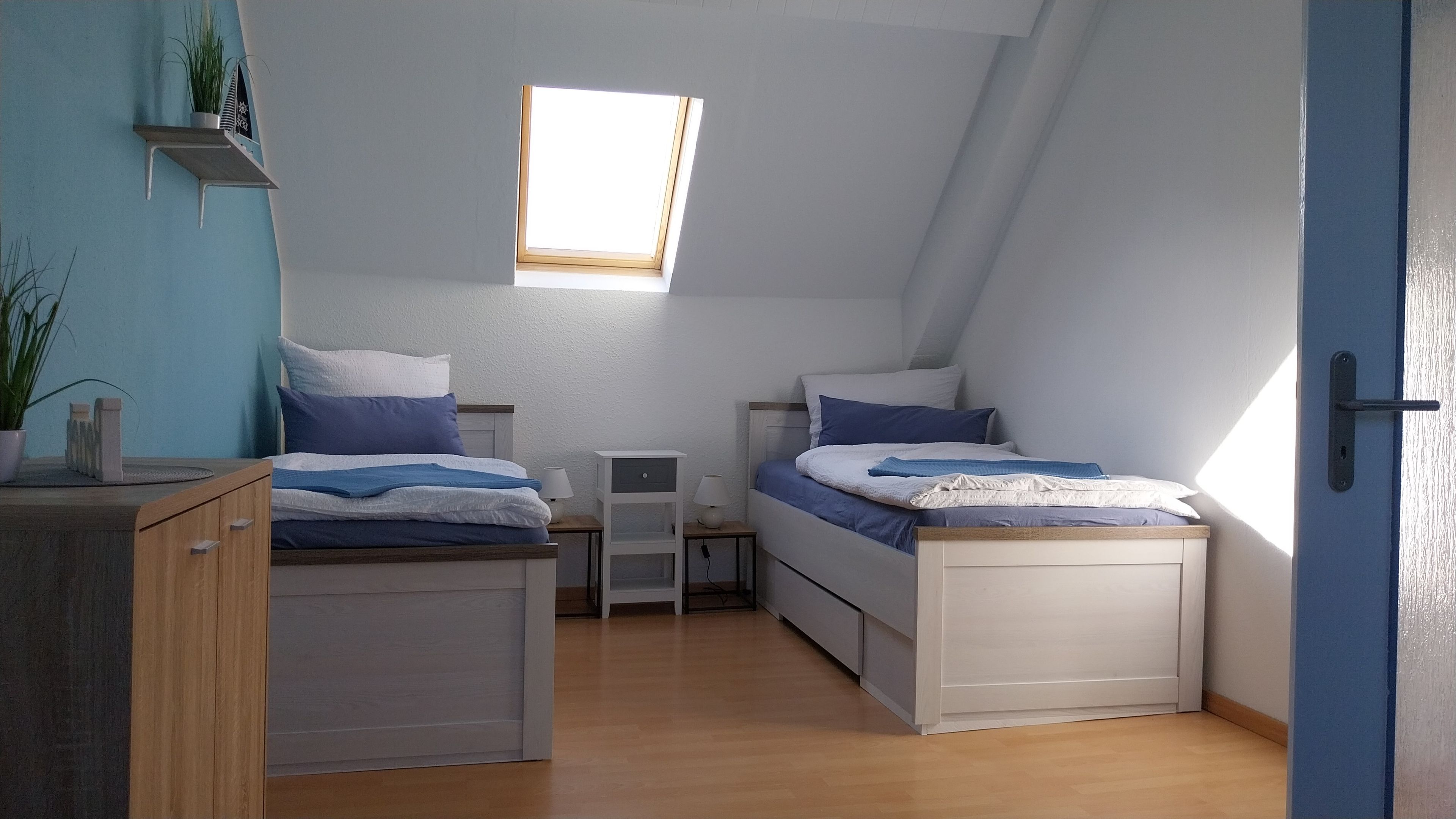 A small, well-lit room with two beds, a desk, a window, and a door.