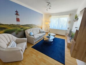 A living room with a sofa, armchair, and coffee table, a painting of a lighthouse, and a window with curtains.