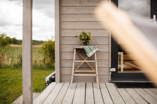 A wooden deck with a small table and a ladder against a building wall, with a garden in the background.