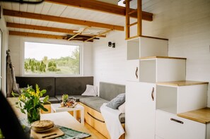 Een knus interieur met een bank, een klein tafeltje met een vaas en een mand, en een raam met uitzicht op groen.