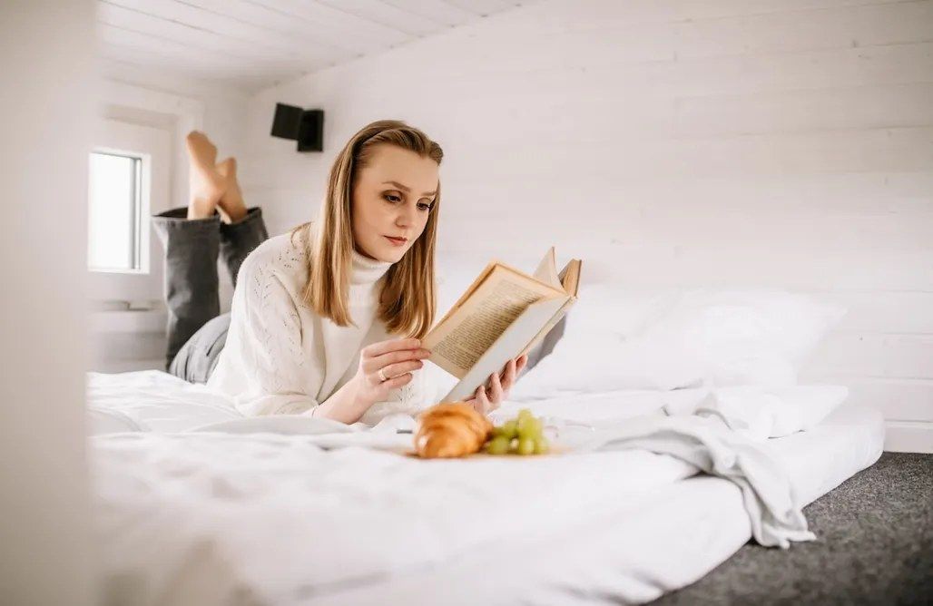A person lying on a bed reading a book with a window and a wall-mounted TV in the background.