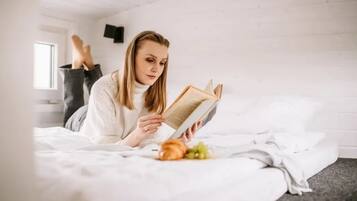 A person lying on a bed reading a book with a window and a wall-mounted TV in the background.