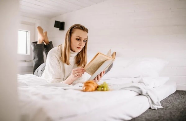 A person lying on a bed reading a book with a window and a wall-mounted TV in the background.