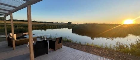 Una terraza de madera con mobiliario exterior, con vistas a una masa de agua tranquila y un atardecer.
