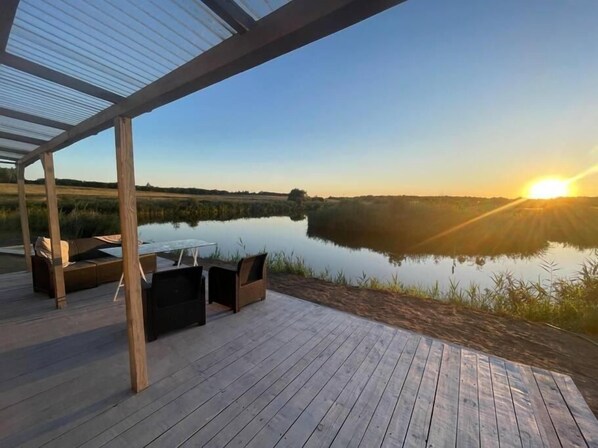 A wooden deck with outdoor seating, overlooking a calm body of water and a sunset.
