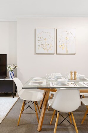 A dining area with a glass table set for four, white chairs, and two framed floral artworks on the wall. - Suite 1108 : Spacious CBD 3 Beds +Pool, Gym & Parking (Brisbane City)