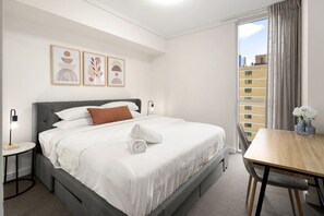 A hotel room with a bed, a bedside table, a chair, a window with curtains, and artwork on the wall. - Central Eco-Luxe 2BR, 2BTH • Pool, Gym & Parking (Brisbane City)