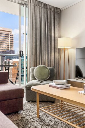 A hotel room with a sofa, a chair, a table, a TV, and a view of the city through large windows. - Central Eco-Luxe 2BR, 2BTH • Pool, Gym & Parking (Brisbane City)