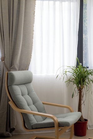 Een moderne fauteuil met een bekleed rugleuning en houten frame, geplaatst naast een potplant met lange groene bladeren, in een kamer met een groot raam en dunne gordijnen.